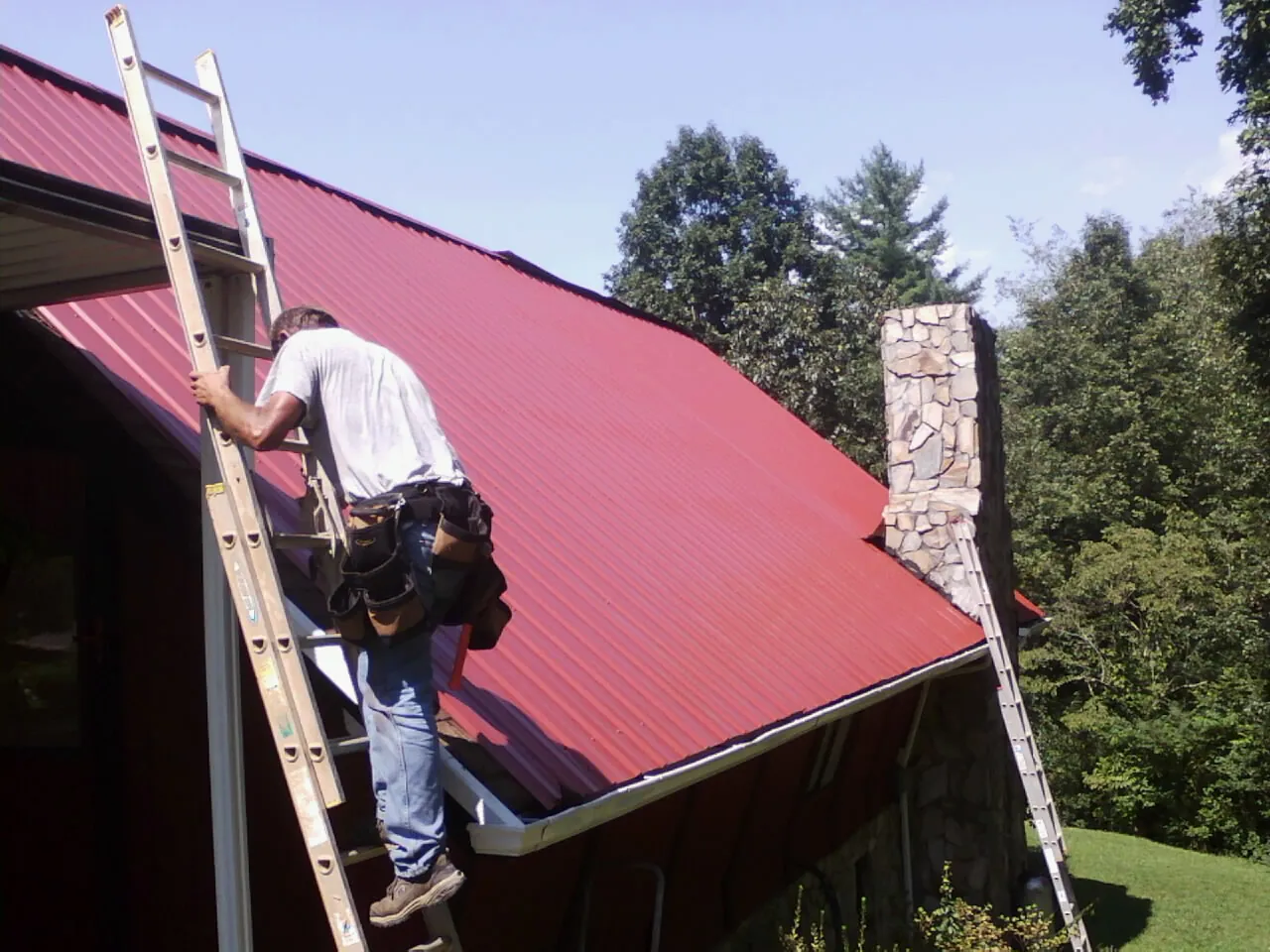 Roofer on a ladder beside a red metal roof and stone chimney — Soffit & Fascia Repair by Roofing Brothers in Cape Neddick