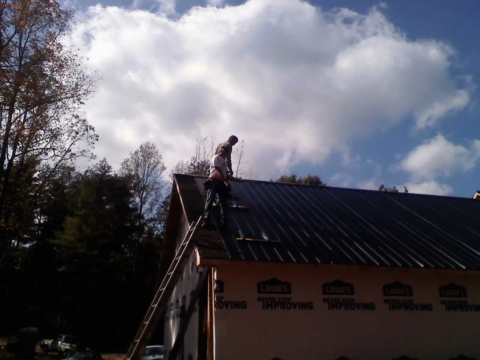 Roofer fastening dark metal panels high against a cloudy sky, from a Roof Inspection job in Cape Neddick